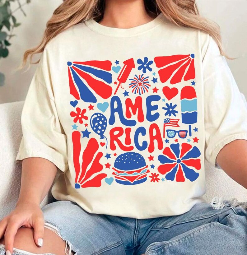 以下が含まれることがあります： 赤、白、青のグラフィックデザインが施された白いTシャツ。グラフィックにはレトロなフォントで「AMERICA」という言葉が書かれており、星、ハート、花火、その他の愛国的なイメージで囲まれています。