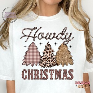 May include: White t-shirt with the text "Howdy Christmas" in brown lettering. The text is surrounded by three Christmas trees, one with a plaid pattern, one with a leopard print, and one with gold glitter.