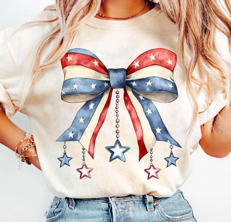 May include: A white t-shirt with a red, white, and blue bow with stars. The bow has a string of beads hanging from it with stars at the end of each string.