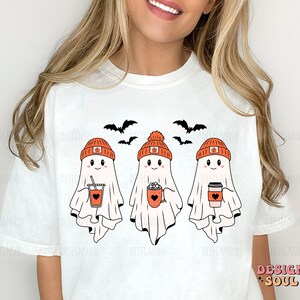 May include: White t-shirt featuring three cartoon ghosts wearing orange pumpkin hats and holding fall-themed drinks. Black bats fly above the ghosts. The text "DESIGN SOUL" is in the lower right corner.