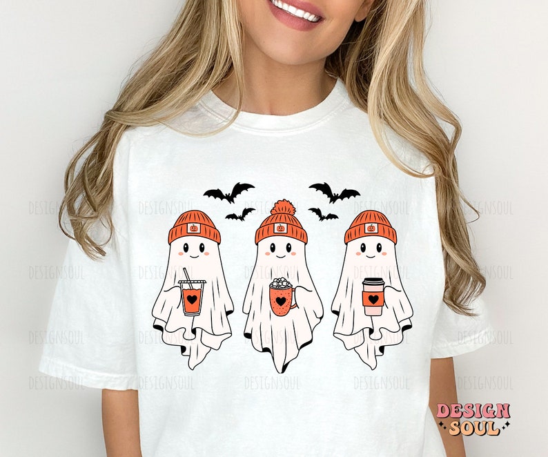 May include: White t-shirt featuring three cartoon ghosts wearing orange pumpkin hats and holding fall-themed drinks. Black bats fly above the ghosts. The text "DESIGN SOUL" is in the lower right corner.
