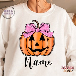 May include: White sweatshirt with a pumpkin design. The pumpkin has a pink bow and a jack-o'-lantern face. The text "Name" is printed below the pumpkin.