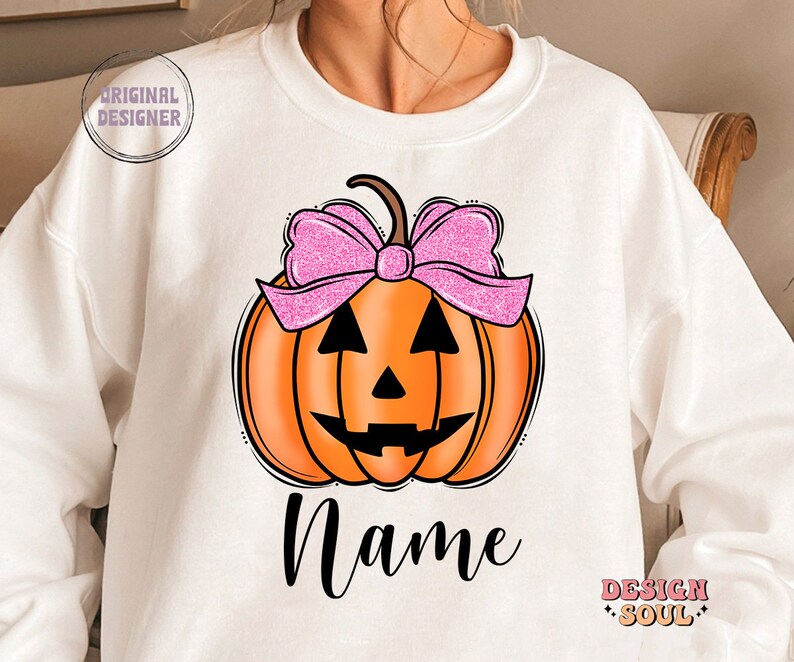 May include: White sweatshirt with a pumpkin design. The pumpkin has a pink bow and a jack-o'-lantern face. The text "Name" is printed below the pumpkin.