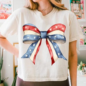 K&ouml;nnte beinhalten: Ein wei&szlig;es T-Shirt mit einem gro&szlig;en rot-wei&szlig;-blauen Bogen, der auf der Vorderseite aufgedruckt ist. Die Schleife besteht aus Stoff mit Sternen und Streifen, die an die amerikanische Flagge erinnern.