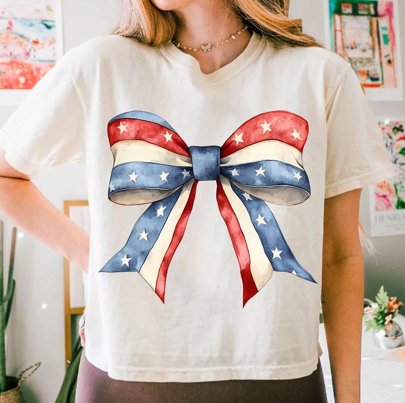 K&ouml;nnte beinhalten: Ein wei&szlig;es T-Shirt mit einem gro&szlig;en rot-wei&szlig;-blauen Bogen, der auf der Vorderseite aufgedruckt ist. Die Schleife besteht aus Stoff mit Sternen und Streifen, die an die amerikanische Flagge erinnern.