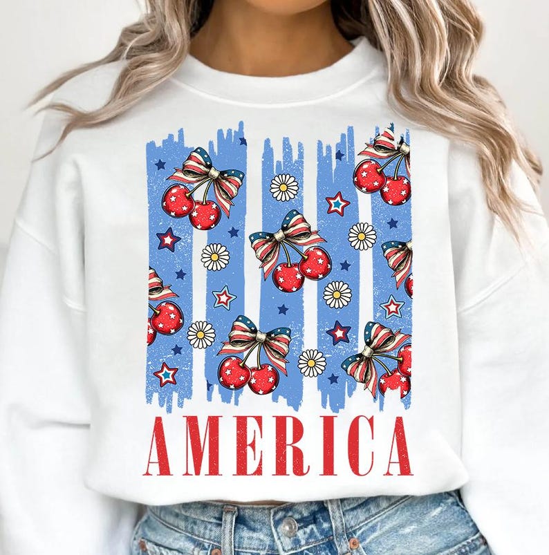 May include: White sweatshirt with a blue and red graphic print of cherries, stars, and daisies. The graphic is a stylized American flag with the word "AMERICA" printed below it.