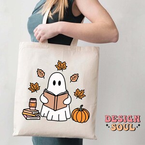 May include: A white tote bag with a ghost reading a book, surrounded by orange leaves and a pumpkin. The text "DESIGN SOUL" is printed on the bottom right corner of the bag.