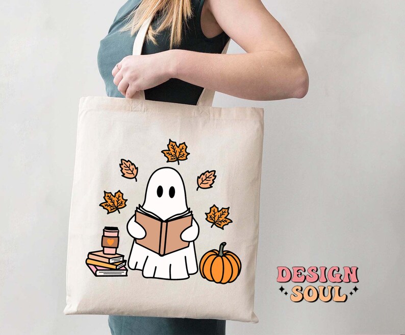 May include: A white tote bag with a ghost reading a book, surrounded by orange leaves and a pumpkin. The text "DESIGN SOUL" is printed on the bottom right corner of the bag.