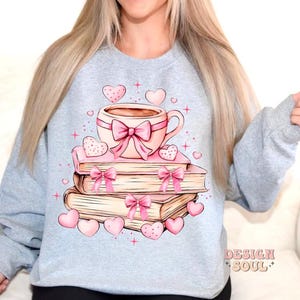 K&ouml;nnte beinhalten: Ein hellgraues Sweatshirt mit einer rosa und wei&szlig;en Illustration einer Tasse Kaffee auf einem Stapel B&uuml;cher mit rosa Schleifen und Herzen.