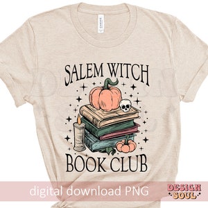 Op de afbeelding: Een beige T-shirt met de tekst "Salem Witch Book Club" gedrukt op de voorkant. Het ontwerp bevat een stapel boeken, een pompoen, een schedel en een kaars.