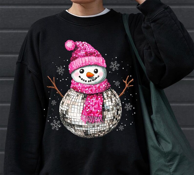 May include: Black sweatshirt with a pink and silver snowman wearing a disco ball as a body. The snowman is wearing a pink hat and scarf with white snowflakes in the background.