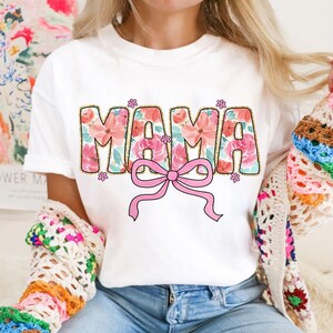May include: White t-shirt with a floral design that spells out "MAMA" with a pink bow in the center.