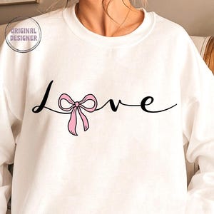 May include: White sweatshirt with the word "Love" written in black cursive with a pink bow in the middle of the word.