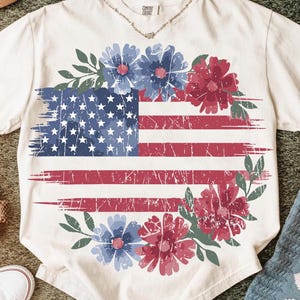 Puede incluir: Camiseta color crema con un dise&ntilde;o desgastado de la bandera estadounidense y detalles florales azules y rojos. Las estrellas y rayas de la bandera se complementan con una corona de flores azules y rojas y hojas verdes, creando un tema patri&oacute;tico y floral.