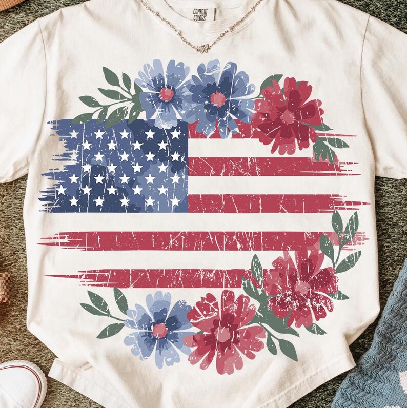 Puede incluir: Camiseta color crema con un dise&ntilde;o desgastado de la bandera estadounidense y detalles florales azules y rojos. Las estrellas y rayas de la bandera se complementan con una corona de flores azules y rojas y hojas verdes, creando un tema patri&oacute;tico y floral.