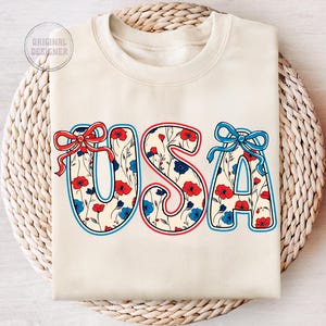 May include: A cream-colored sweatshirt with the word "USA" printed on the front. The letters are outlined in red and blue and filled with a pattern of red, white, and blue flowers.