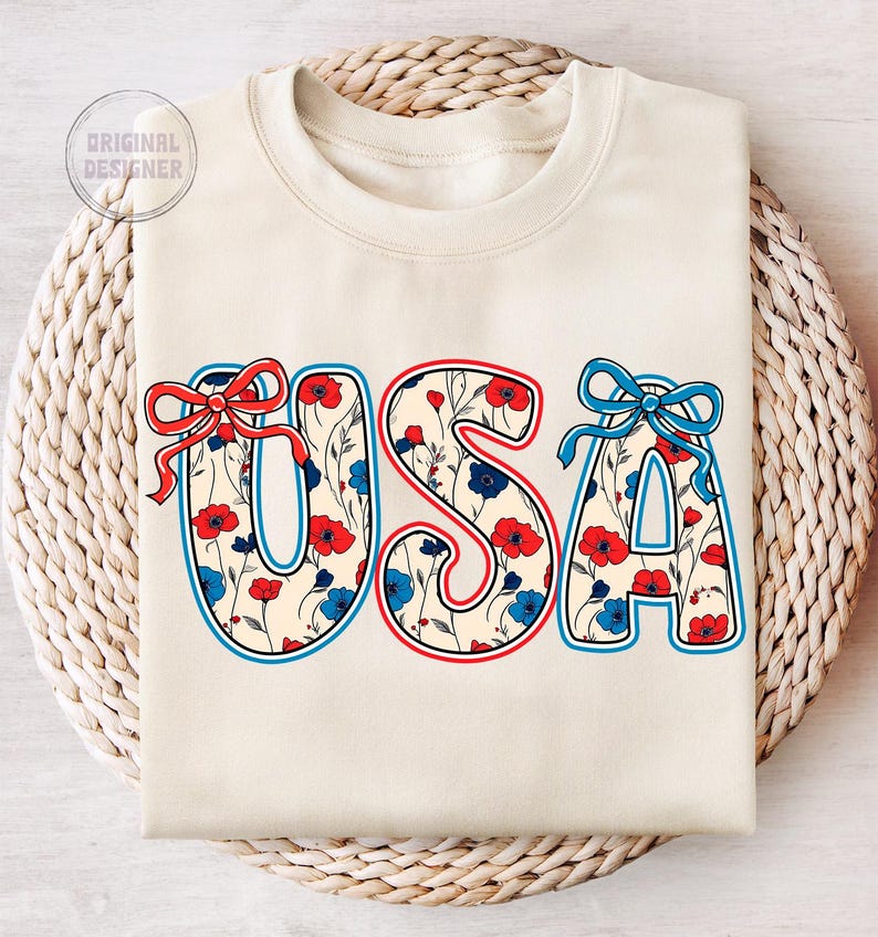 May include: A cream-colored sweatshirt with the word "USA" printed on the front. The letters are outlined in red and blue and filled with a pattern of red, white, and blue flowers.