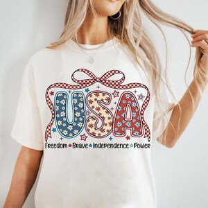 May include: White t-shirt with a red, white, and blue USA graphic. The graphic features a bow, stars, and flowers. The text "Freedom * Brave * Independence * Power" is printed below the graphic.