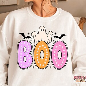 Op de afbeelding: Wit sweatshirt met een roze, oranje en witte "BOO" grafiek met een geest en vleermuizen. De tekst is zwart omlijnd.