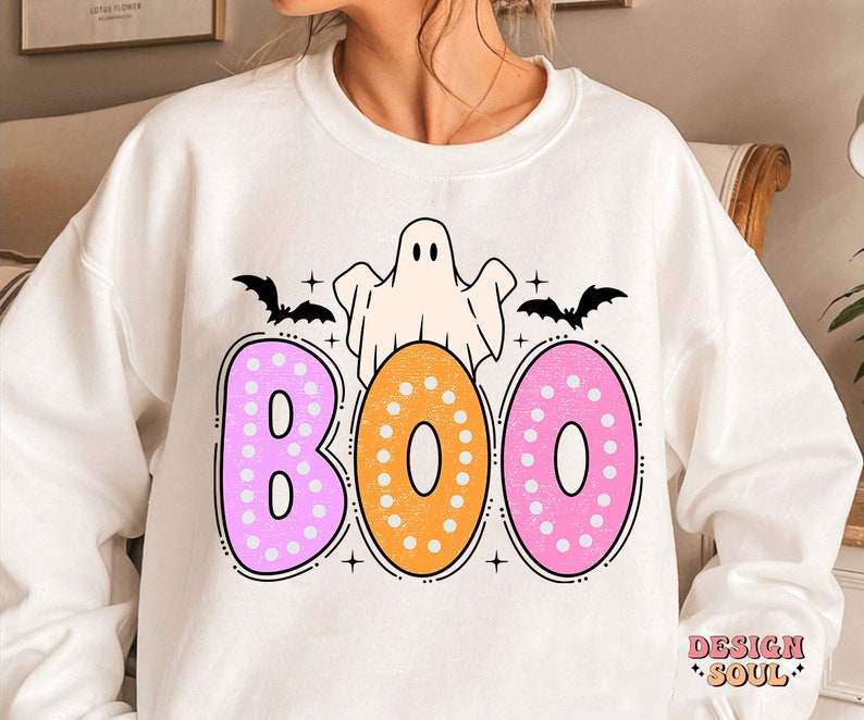 Op de afbeelding: Wit sweatshirt met een roze, oranje en witte "BOO" grafiek met een geest en vleermuizen. De tekst is zwart omlijnd.