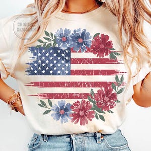 Puede incluir: Camiseta color crema con un dise&ntilde;o desgastado de la bandera estadounidense, detalles florales rojos y azules, y una corona floral. El texto "ORIGINAL DESIGNER" es visible. El dise&ntilde;o evoca una est&eacute;tica vintage.