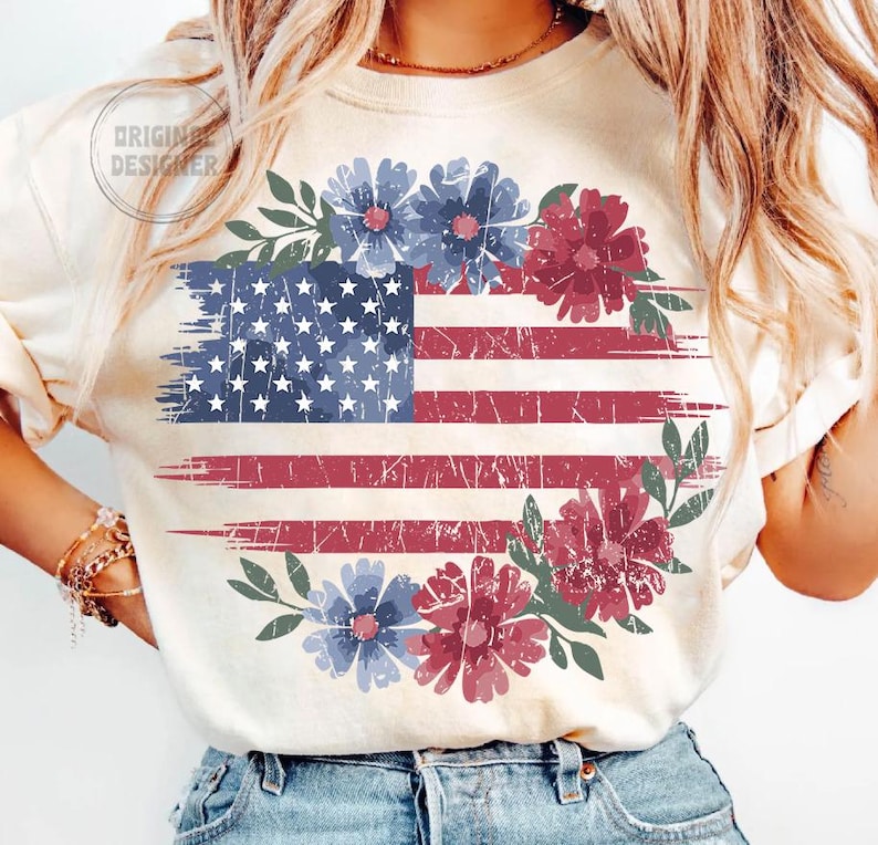 Puede incluir: Camiseta color crema con un dise&ntilde;o desgastado de la bandera estadounidense, detalles florales rojos y azules, y una corona floral. El texto "ORIGINAL DESIGNER" es visible. El dise&ntilde;o evoca una est&eacute;tica vintage.
