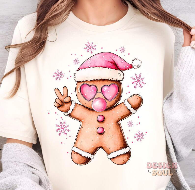 May include: A white t-shirt with a gingerbread man wearing a pink Santa hat and pink heart-shaped sunglasses. The gingerbread man is holding up two fingers in a peace sign and has a pink bubble coming out of his mouth. The background is white with pink snowflakes.