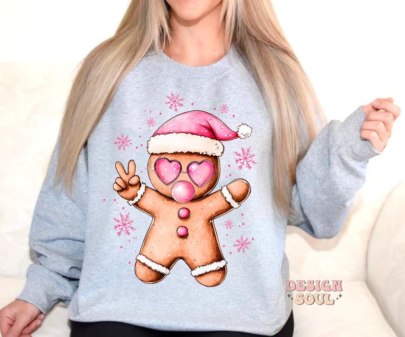 May include: A light blue sweatshirt with a gingerbread man wearing a pink Santa hat and pink sunglasses. The gingerbread man is holding up two fingers in a peace sign. The sweatshirt has a snowflake pattern and the text "Design Soul" on the bottom right corner.