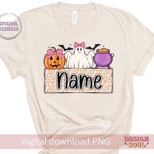 May include: A beige t-shirt with a Halloween-themed design featuring a ghost, a pumpkin, and a cauldron. The design includes the text "Name" in a black font.