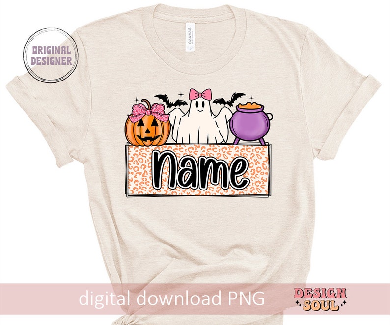 May include: A beige t-shirt with a Halloween-themed design featuring a ghost, a pumpkin, and a cauldron. The design includes the text "Name" in a black font.