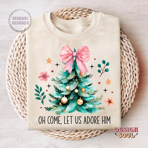 May include: A beige sweatshirt with a Christmas tree design. The tree is green with gold ornaments and a pink bow. The text "Oh come, let us adore Him" is printed on the sweatshirt.
