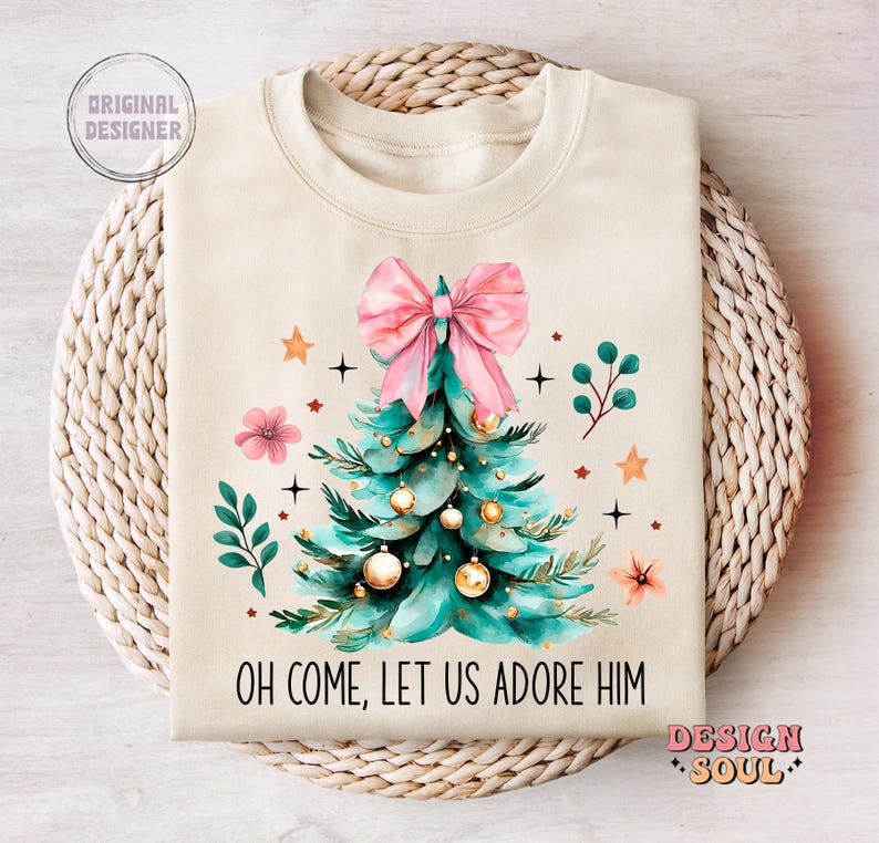 May include: A beige sweatshirt with a Christmas tree design. The tree is green with gold ornaments and a pink bow. The text "Oh come, let us adore Him" is printed on the sweatshirt.