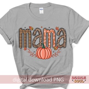May include: A gray t-shirt with the word "mama" in a fall-themed design. The letters are decorated with plaid, leopard print, and fall leaves. A pumpkin is in the center of the design.