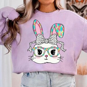 May include: A lavender sweatshirt with a white bunny wearing glasses and a black and white polka dot bow. The bunny's ears are pink and blue with a colorful pattern.
