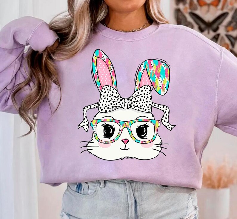 May include: A lavender sweatshirt with a white bunny wearing glasses and a black and white polka dot bow. The bunny's ears are pink and blue with a colorful pattern.