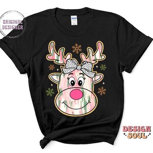 May include: Black t-shirt with a pink and green reindeer face graphic. The reindeer has a pink nose and a pink bow with white polka dots. The graphic is surrounded by snowflakes.