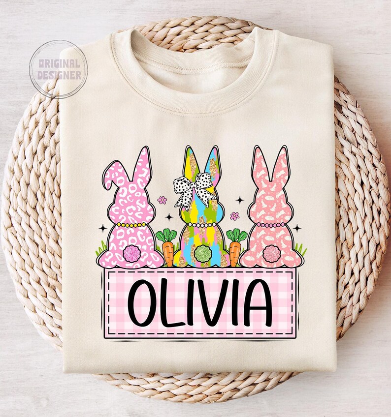 May include: A beige sweatshirt with a pink and white checkered banner that says "OLIVIA". Three pink, yellow, and blue bunny rabbits are sitting on top of the banner. The bunnies have glitter and a bow on their heads.