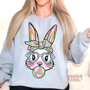 May include: A light grey sweatshirt featuring a colourful cartoon bunny wearing heart-shaped sunglasses and a polka dot headband. The bunny is holding a colourful lollipop in its mouth.