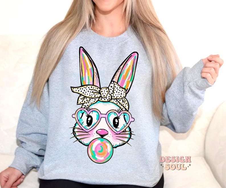 May include: A light grey sweatshirt featuring a colourful cartoon bunny wearing heart-shaped sunglasses and a polka dot headband. The bunny is holding a colourful lollipop in its mouth.