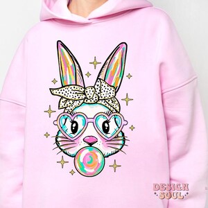 May include: Pink hoodie with a graphic of a cartoon bunny wearing sunglasses and a headband. The bunny is holding a colorful lollipop. The hoodie has the text "DESIGN SOUL" on the bottom right corner.