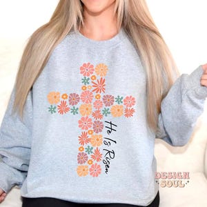 May include: A light gray sweatshirt with a floral cross design and the text "He is Risen".