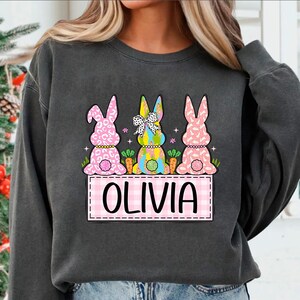 May include: A gray sweatshirt with a pink, blue, and white bunny rabbit design. The bunnies are wearing bows and have carrots in front of them. The text "Olivia" is written in pink on a pink and white checkered background.
