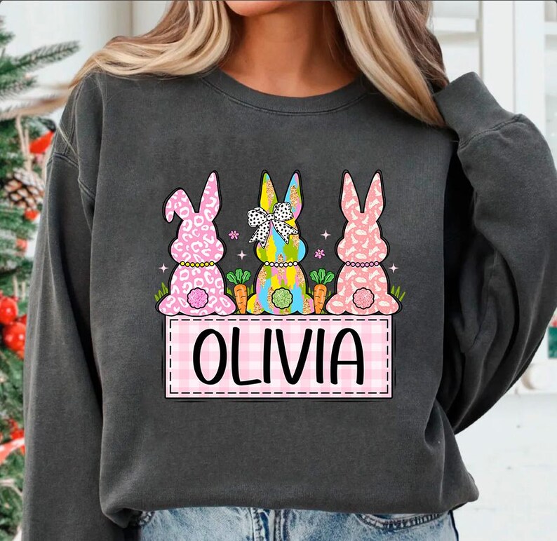 May include: A gray sweatshirt with a pink, blue, and white bunny rabbit design. The bunnies are wearing bows and have carrots in front of them. The text "Olivia" is written in pink on a pink and white checkered background.