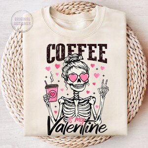 May include: A beige sweatshirt with the text "COFFEE is my VALENTINE" in black. A cartoon skeleton wearing pink sunglasses and a bun holds a pink and white coffee cup. Pink hearts are scattered around the skeleton.