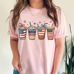 May include: Pink t-shirt with a graphic of four iced coffee cups decorated with the American flag.