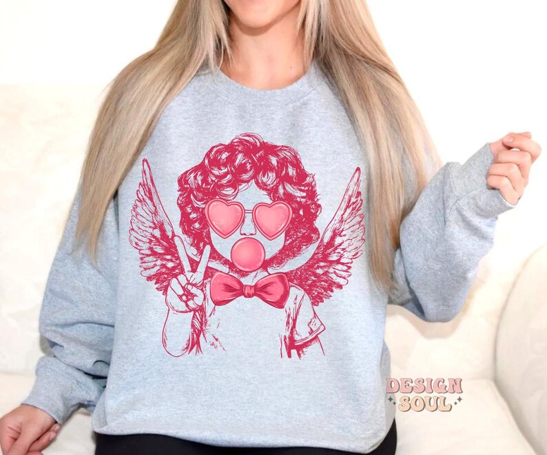 May include: A grey sweatshirt featuring a pink graphic of a cherub with angel wings, heart-shaped sunglasses, and a pink bubble. The cherub is giving the peace sign.