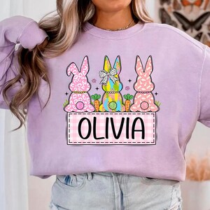 May include: A lavender sweatshirt with a graphic of three Easter bunnies. The bunnies are wearing colorful clothes and holding carrots. The text "Olivia" is written in a pink and white checkered box.