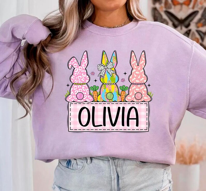 May include: A lavender sweatshirt with a graphic of three Easter bunnies. The bunnies are wearing colorful clothes and holding carrots. The text "Olivia" is written in a pink and white checkered box.