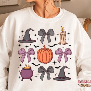 Puede incluir: Sudadera blanca con un dise&ntilde;o de tem&aacute;tica de Halloween que presenta una calabaza, sombreros de bruja, lazos, un caldero y una vela. El dise&ntilde;o est&aacute; en tonos de morado, naranja y gris.