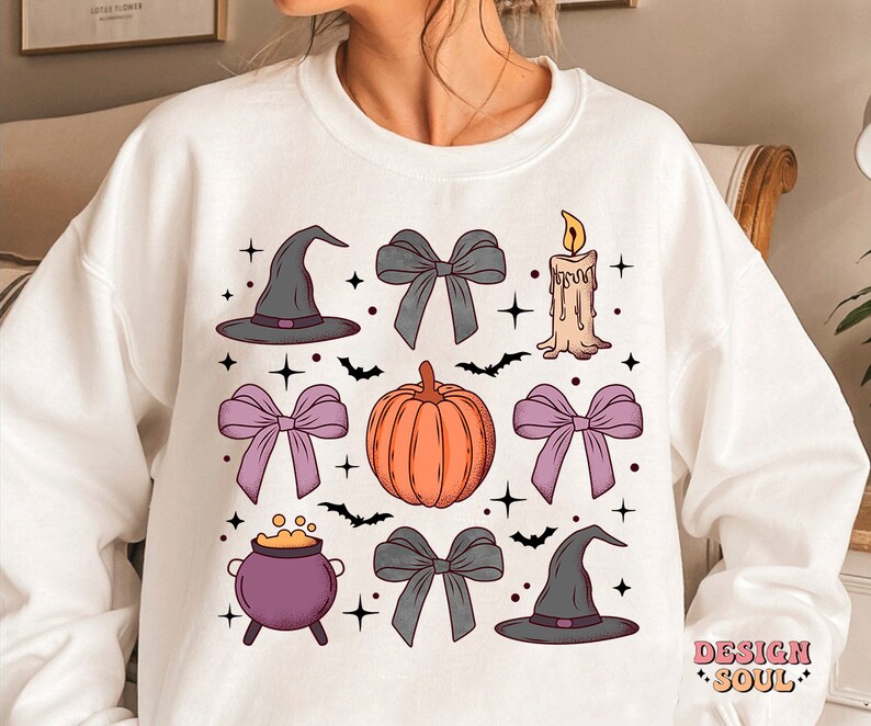 Puede incluir: Sudadera blanca con un dise&ntilde;o de tem&aacute;tica de Halloween que presenta una calabaza, sombreros de bruja, lazos, un caldero y una vela. El dise&ntilde;o est&aacute; en tonos de morado, naranja y gris.